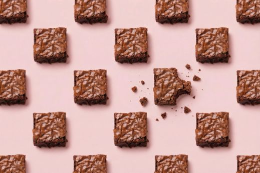Pattern of chocolate brownies on a pink background with one bitten piece and scattered crumbs.